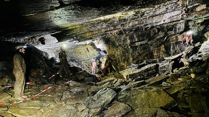 Mehrere Menschen stehen in schlammigen Schutzanzügen und mit Stirnlampen in einer dunklen Höhle