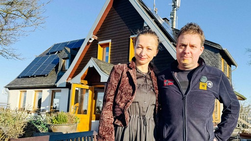 Thomas und Aleksandra Prinz, ein Mann und eine Frau mittleren Alters, stehen vor einem Haus mit gelben Fesnsteröäden und Türrahmen. Am linken Bildrand sind Zweige eines Baums zu sehen und auf dem Dach des Hauses sind Solarpanele angebracht.