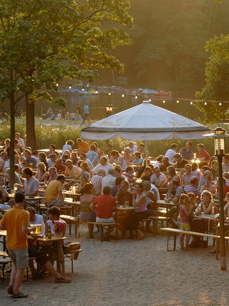 Menschen sitzen in der Abendsonne in einem Biergarten inmitten eines Parks