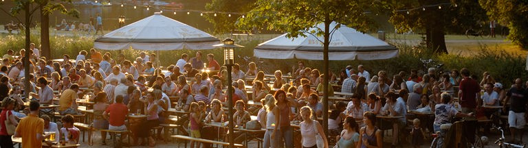 Menschen sitzen in der Abendsonne in einem Biergarten inmitten eines Parks