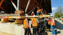 Mehrere Personen in orangenen Warnwesten und mit weißen Helmen auf einem hölzernen Gerüst