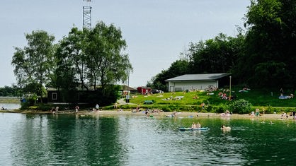 Menschen schwimmen entweder im Wasser oder sonnen sich auf einer Wiese am Ufer.