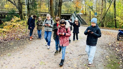 Eine Gruppe an Kindern begleitet von einer Lehrerin läuft durch den Wald. Alle ausgerüstet mit einem Handy oder Tablet und der App. 