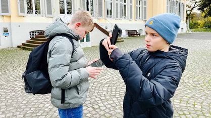 Zwei Jungs stehen mit einem Handy und einem Tablet vor dem Schloss Morsbroich