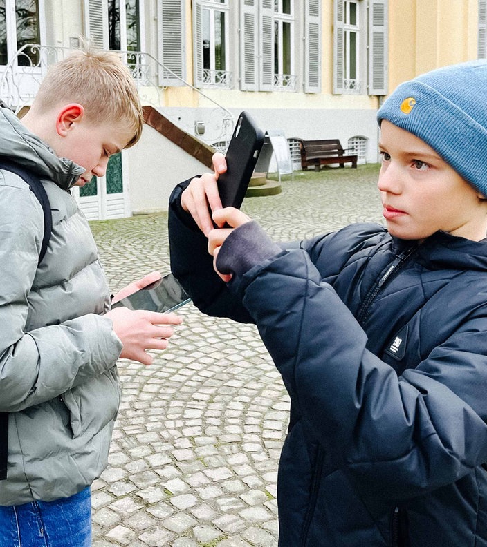 Zwei Jungs stehen mit einem Handy und einem Tablet vor dem Schloss Morsbroich