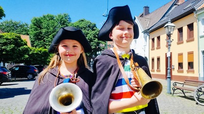 Ein Mädchen und ein Junge stehen mit Umhang, Mütze und Hörnern in einer Altstadt.