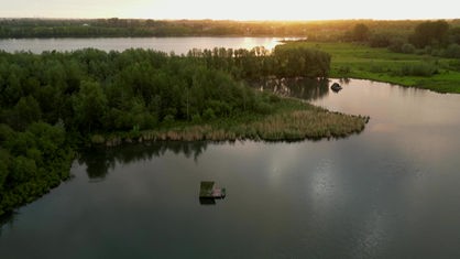 Auf einem See schwimmt ein Floß. Auf der Verande sitzen zwei Menschen auf Campingstühlen.