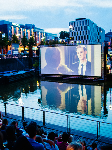 Eine Szene des Open-Air-Kinos am Kölner Rheinauhafen, mit dem Hafen im Vordergrund und dem Kinoareal im Hintergrund.