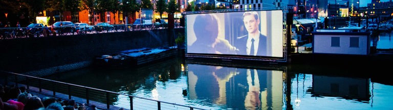 Eine Szene des Open-Air-Kinos am Kölner Rheinauhafen, mit dem Hafen im Vordergrund und dem Kinoareal im Hintergrund.