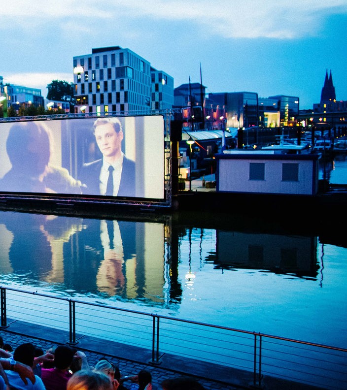 Eine Szene des Open-Air-Kinos am Kölner Rheinauhafen, mit dem Hafen im Vordergrund und dem Kinoareal im Hintergrund.