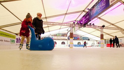 Ein Kind fährt auf einer Eisbahn Schlittschuh, es hält sich an einem blauen Wal als Schiebetier fest, daneben ist eine Erwachsene