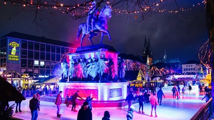 eine Eisbahn mit bunter Beleuchtung, die um ein Reiterdenkmal führt, im Hintergrund ist der Kölner Dom zu sehen