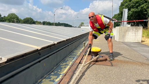 Ein Mann in gelber Arbeitskleidung mit Rettungsweste und Sonnenbrille bückt sich und greift nach einem Schiffstau