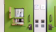 Frauenforensik der LVR-Klinik Bedburg-Hau: Eine grüne Zimmertür mit geöffneten Klappen. Dahinter eine Frau, die aus einem vergitterten Fenster blickt