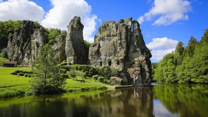 Die graue Felsformation der Externsteine, dahinter ein blauer Himmel, im Vordergrund ein Teich