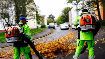 Mert Aybey und sein Kollege sind mit Laubbläsern an einer Straße in einem Wohngebiet unterwegs.