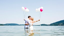 Ein Hochzeitspaar steht im Wasser und küsst sich. Sie halten pinke und weiße Luftballons.