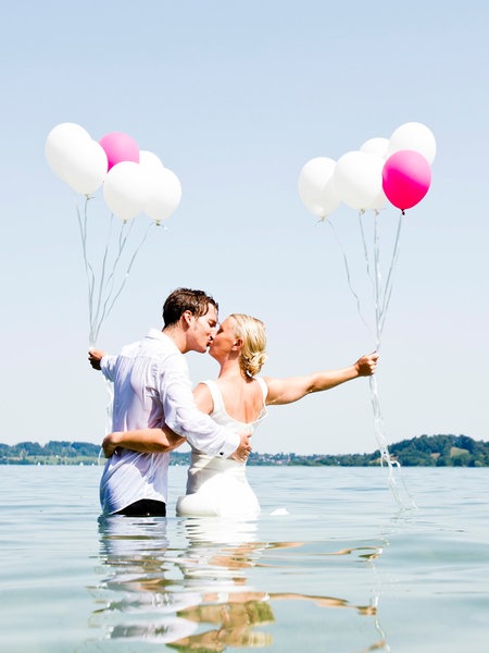 Ein Hochzeitspaar steht im Wasser und küsst sich. Sie halten pinke und weiße Luftballons.