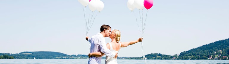 Ein Hochzeitspaar steht im Wasser und küsst sich. Sie halten pinke und weiße Luftballons.