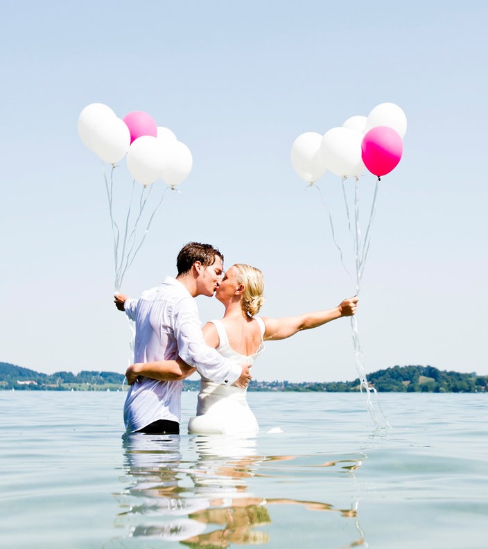 Ein Hochzeitspaar steht im Wasser und küsst sich. Sie halten pinke und weiße Luftballons.