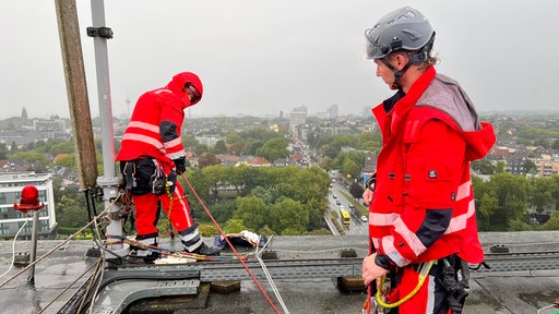 Zwei industriekletterer stehen in roten Anzügen und mit Seilen gesichert auf der Spitze eines Hochhauses in Essen, im Hintergrund ist die Stadt von oben zu sehen