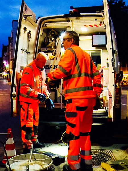 Zwei Männer in orangener Arbeitskleidung stehen mit ihrem Auto vor einem Kanalschacht.