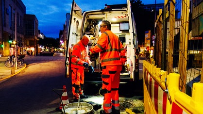 Zwei Männer in orangener Arbeitskleidung stehen mit ihrem Auto vor einem Kanalschacht.