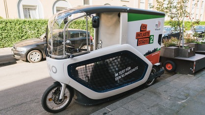 Ein großes Lastenrad mit viel Stauraum und einem Dach steht an einer Straße.