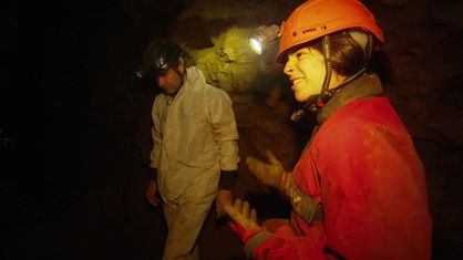 In einer dunklen Höhle steht eine Tourguide und erklärt den Teilnehmern worauf sie jetzt achten müssen. Sie trägt einen roten Overall mit rotem Helm und Stirnlampe.