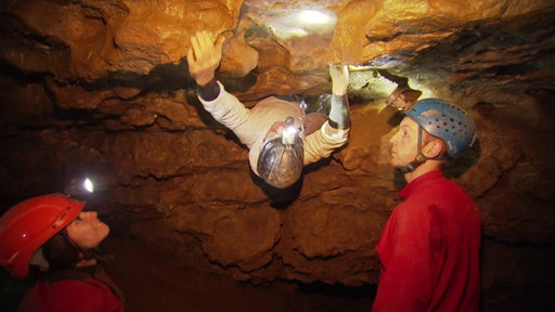 Zwei Tourguides in roten Overalls stehen unterhalb einer Felsspalte. Von oben klettert ein Teilnehmer in weißem Overall aus der Spalte.