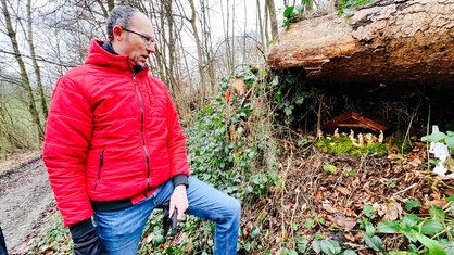 Ein Mann mit einer roten Jacke steht im Wald, am Hang ist eine Krippe aufgebaut