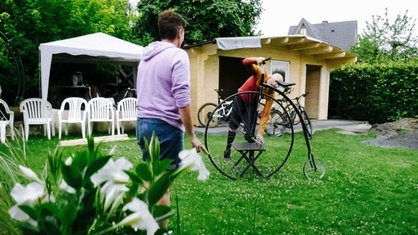 Klaus Lenfort schraubt im Garten an seinem Hochrad und seine Frau schaut ihm zu.