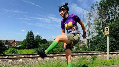 Eine junge Frau gibt auf dem Longboard, sie trägt einen Helm mit Hupe, ein lila Shirt und grüne Strümpfe.