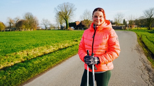 Janine Marx steht in leuchtender Jacke und mit Walking-Stöcken auf einem Weg und blickt in die Kamera.