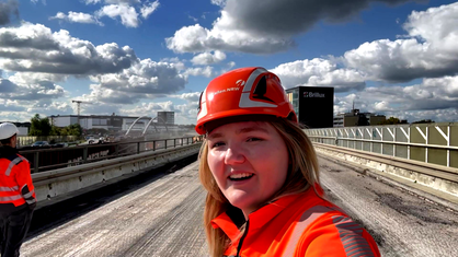 Eine Frau in orangener Jacke und mit Helm steht auf einer Brücke 