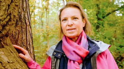 Eine Frau mittleren Alters mit blonden Haaren und einem rosa Schaal schaut in Richtung Kamera. Daneben ein Baum