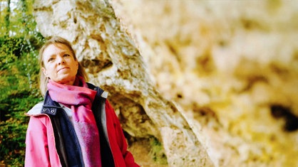 Ein Frau mit blonden Haaren vor einer Felswand. Sie trägt einen rosa Schaal und eine rosa Jacke