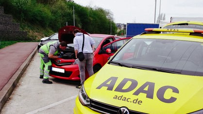 Im Vordergrund ein gelbes Auto mit der schwarzen Aufschrift ADAC auf der Motorhaube, im Hintergrund guckt Pannenhelfer Stefan Sonnemann in den Motorraum eines roten Kleinwagens, die Fahrerin beobachtet ihn dabei