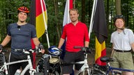 Drei Menschen stehen vor ihren Fahrrädern. Hinter ihnen steht die deutsche, niederländische und belgische Flagge.