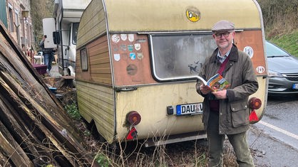 Ralf Kramp vor einem Wohnwagen, in der Hand ein Buch