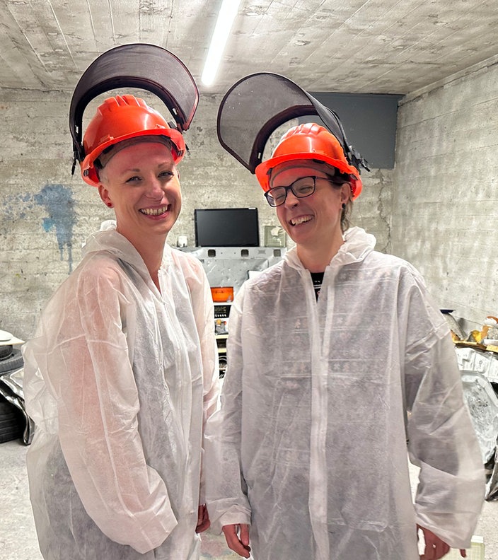 Zwei Frauen stehen in weißen Overalls und mit orangenen Helmen in einem fensterlosen Raum in einem Wutraum, um sie herum steht Schrott