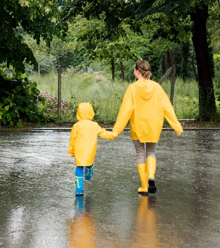 Ein kleines Kind und eine junge Frau laufen Hand in Hand mit Regenmänteln durch den Regen