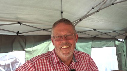Ein Mann in weiß-rot kariertem Hemd steht unter einem Zelt. Er lächelt breit grinsend.