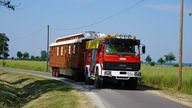 Ein zwanzig Meter langer Zirkuswagen mit einer Feuerwehrzugmaschine fährt zwischen Wiesen auf einer schmalen Straße.