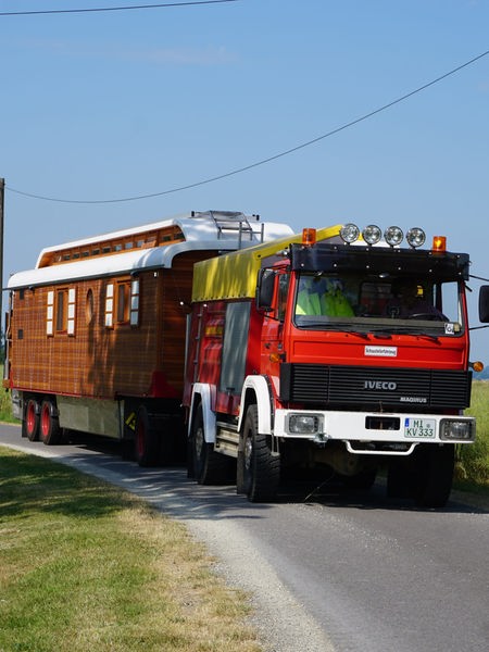 Ein zwanzig Meter langer Zirkuswagen mit einer Feuerwehrzugmaschine fährt zwischen Wiesen auf einer schmalen Straße.