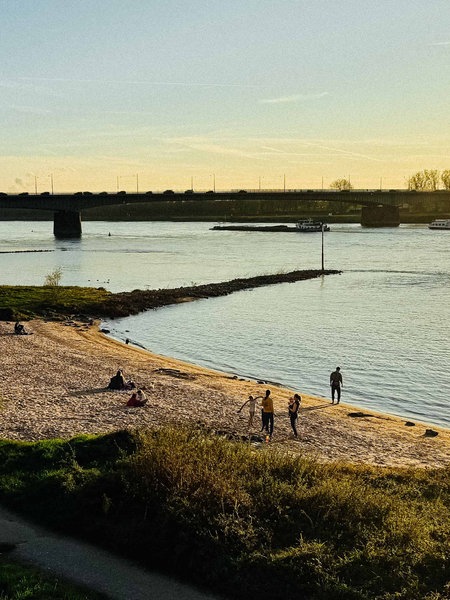 Die Sonne geht an den Sandbuchten am Rhein unter, ein paar Leute sind am Strand und zwei Schiffe fahren über den Rhein entlang