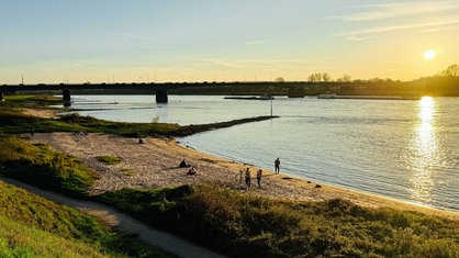 Die Sonne geht an den Sandbuchten am Rhein unter, ein paar Leute sind am Strand und zwei Schiffe fahren über den Rhein entlang