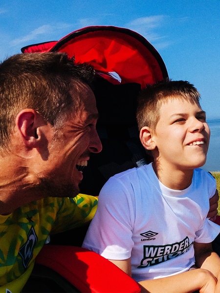 Daniel Buschatz und sein Sohn Niklas am Meer. Daniel kniet neben dem Rollstuhl von Niklas und lächelt seinen Sohn an. Niklas lacht und schaut geradeaus.