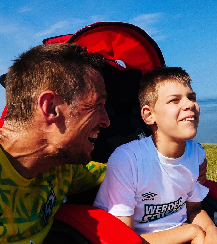 Daniel Buschatz und sein Sohn Niklas am Meer. Daniel kniet neben dem Rollstuhl von Niklas und lächelt seinen Sohn an. Niklas lacht und schaut geradeaus.