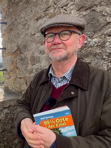 Ralf Kramp vor einer Mauer und hält sein Buch in der Hand
